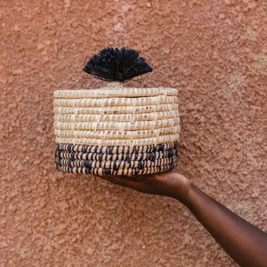 JustOne's natural basket with black details on bottom and lid with a black pompom handle, handcrafted in Uganda