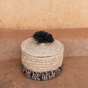 JustOne's natural basket with black details on bottom and lid with a black pompom handle, handcrafted in Uganda
