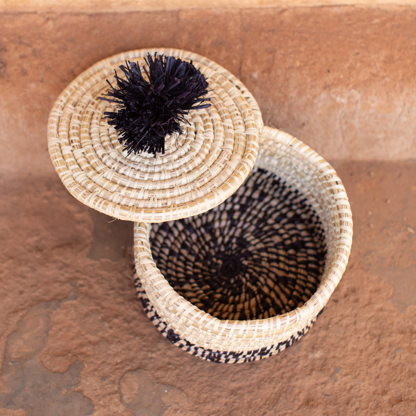 JustOne's natural basket with black details on bottom and lid with a black pompom handle, handcrafted in Uganda