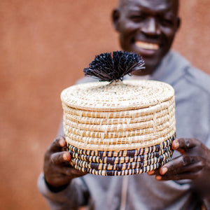 JustOne's natural basket with black details on bottom and lid with a black pompom handle, handcrafted in Uganda