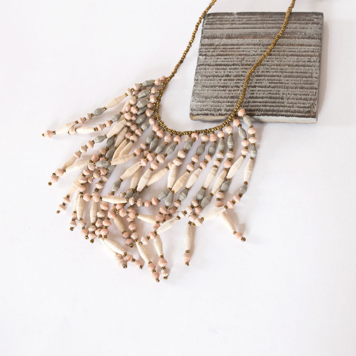 JustOne's necklace with several dangling strings of paper beads in muted grey, pink, and tan colours, handcrafted in Uganda